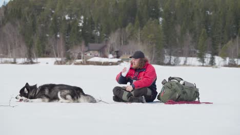 backpacker drinking water while resting with his alaskan malamute dog in frozen landscape