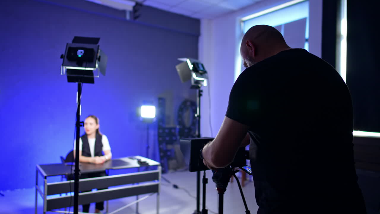Male photographer wearing black t-shirt stands at the camera on tripod setting the equipment for footage in modern studio. Unrecognized girl sits at the desk at blurred backdrop.
