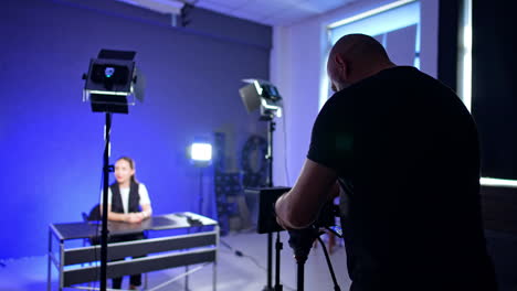 male photographer wearing black t-shirt stands at the camera on tripod setting the equipment for footage in modern studio. unrecognized girl sits at the desk at blurred backdrop.