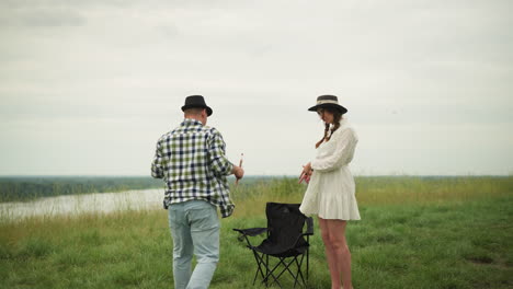 a painter in a checked shirt holds brushes and walks toward a woman in a white dress standing in a grassy field. he sits on a chair in front of her, crossing his legs