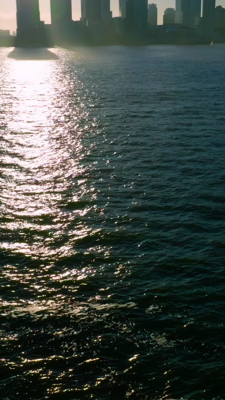 Dark silhouettes of skyscrapers at Manhattan Island. Sun reflecting in the dark water of Hudson river. Vertical video