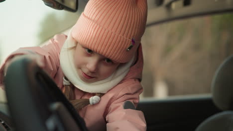 a young girl, dressed in a pink beanie and jacket, sits eagerly in the front passenger seat of a car, playfully turning the steering wheel with excitement. in the background, a man, likely her father