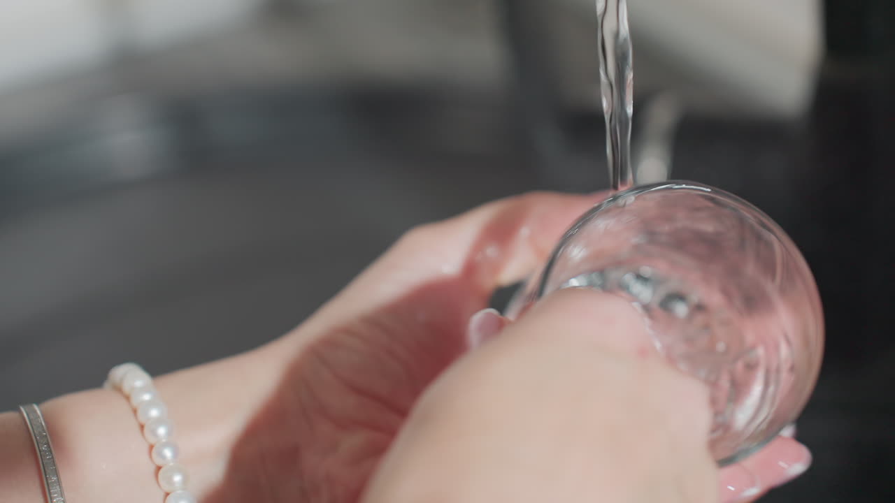 Close up of woman wearing beads thoroughly rinsing glass cup under flowing water at kitchen sink with light reflecting on hand showing careful attention to cleanliness hygiene
