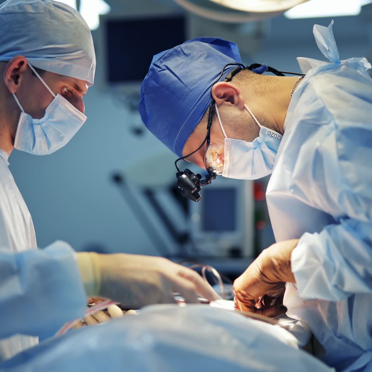 Surgery in clinic. Professional surgeon in medical mask and flashlight on head performing an operation on a patient. Male assistant helps the doctor in the operating room.