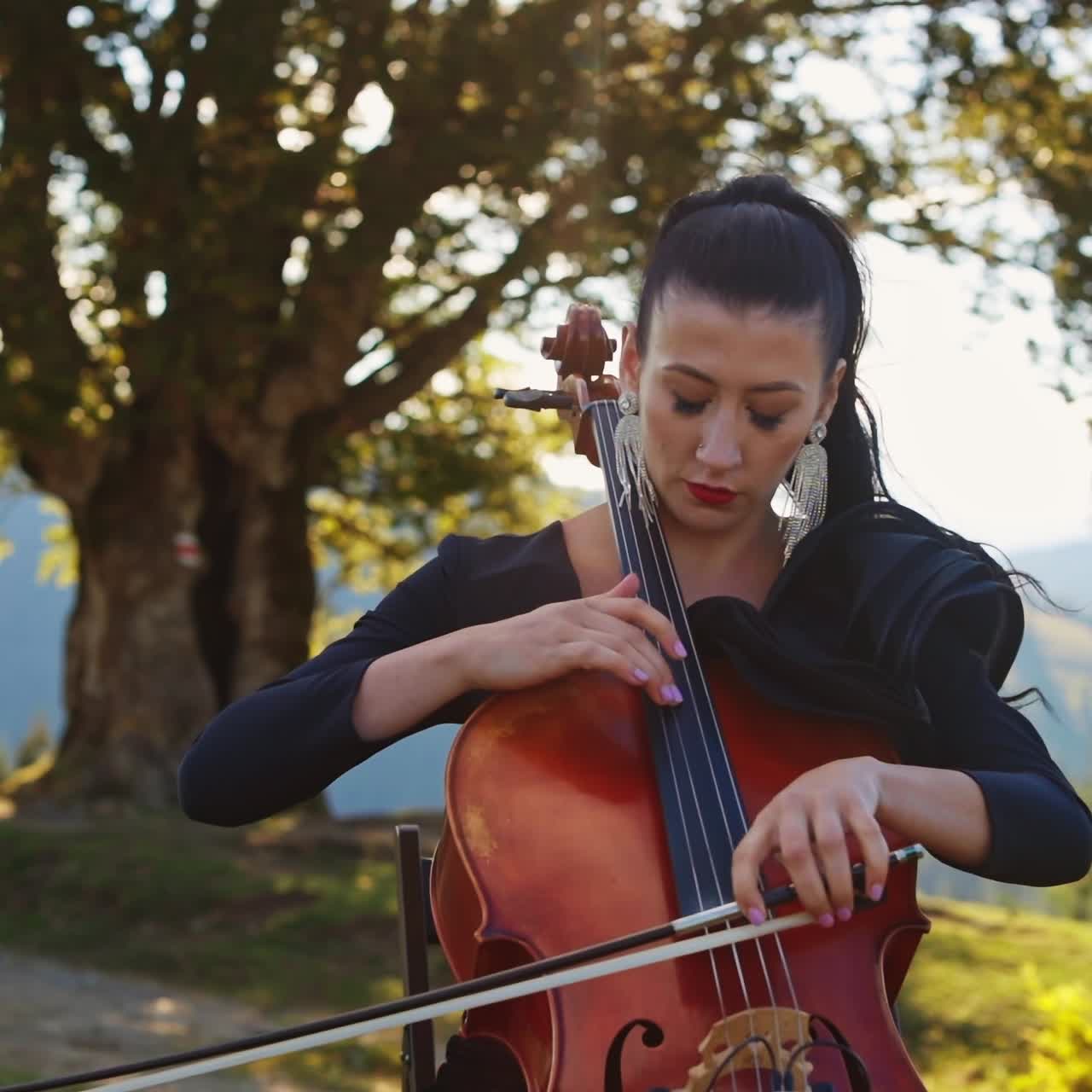 Dark-haired female musician focused on playing cello. Middle-aged woman in black dress plays instrument in the nature backdrop