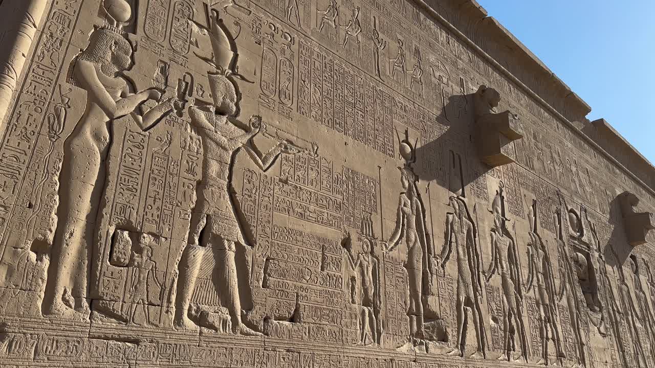 Elokuvamainen panoraamakuva Denderassa sijaitsevan Hathorin temppelin ulkoisesta kivimuurista, jossa on yksityiskohtaisia muinaisen Egyptin faaraoiden ja jumalien reliefejä kirkkaassa päivänvalossa.