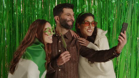 fotografía de estudio de amigos disfrazándose con novedades irlandesas y accesorios posando para selfies celebrando el día de san patricio contra un fondo de oropel verde