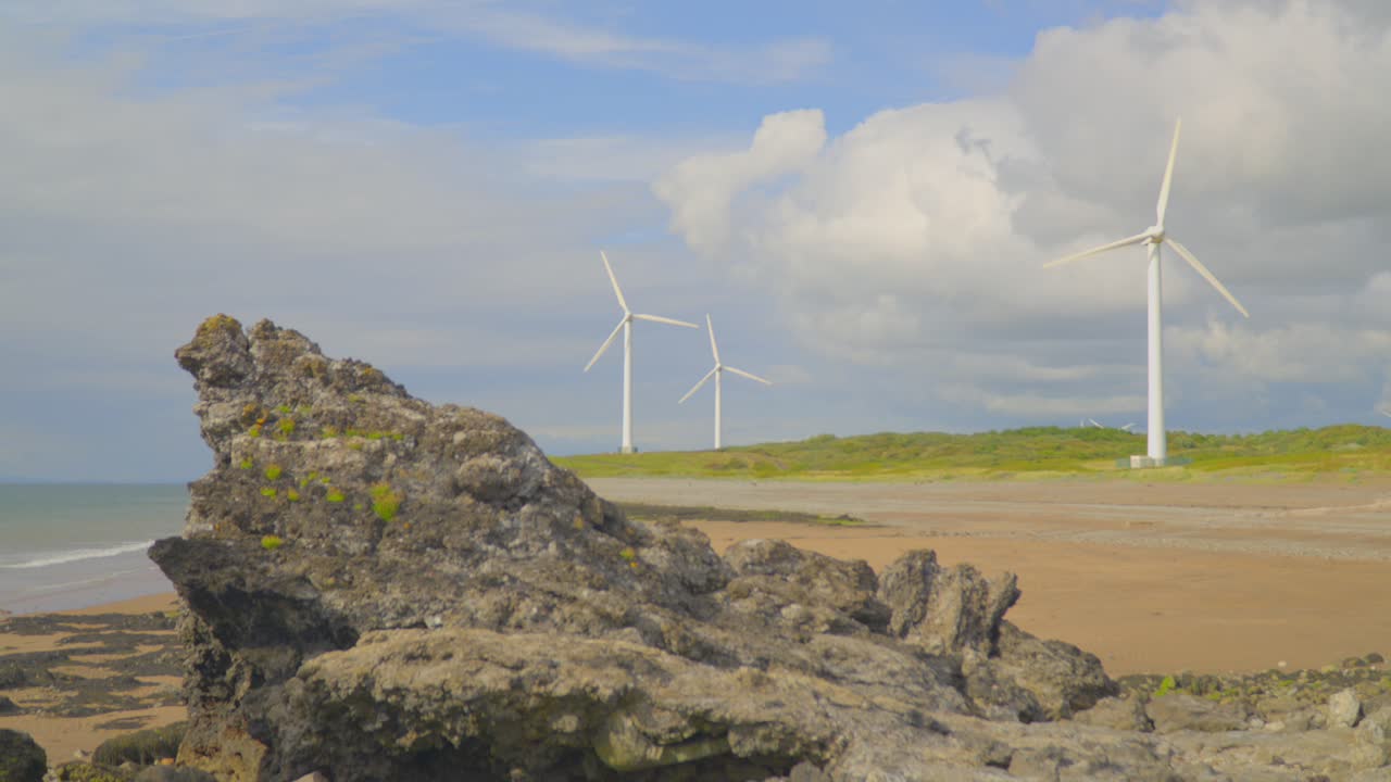 전면의 바위 형성이 있는 구름이 많은 여름날 해변에 있는 풍력 터빈