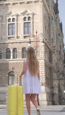 a young beautiful girl with a yellow suitcase is walking on a city street from behind. back view of a girl traveler walking and pulling a suitcase