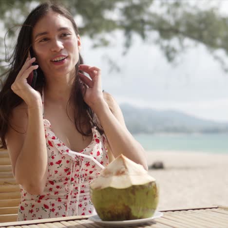 Cheerful-woman-with-coconut-talking-on-phone-on-seaside