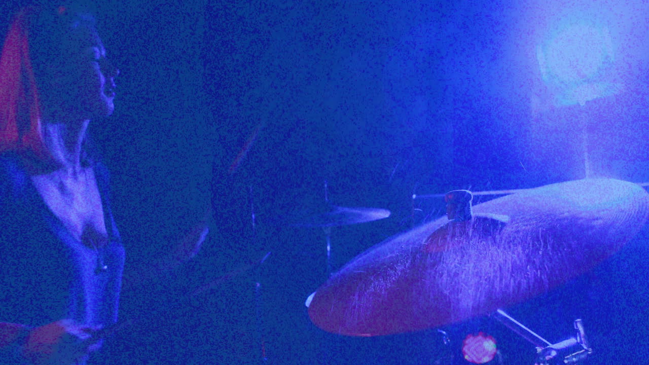 Baterista feminina tocando com baquetas em palco com pouca luz, com notas musicais animadas e luzes neon.