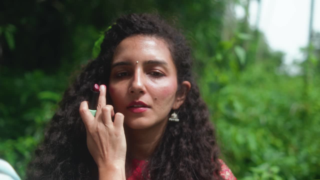 A woman with curly hair gets blush applied on her cheeks by another woman, in front of dense green vegetation