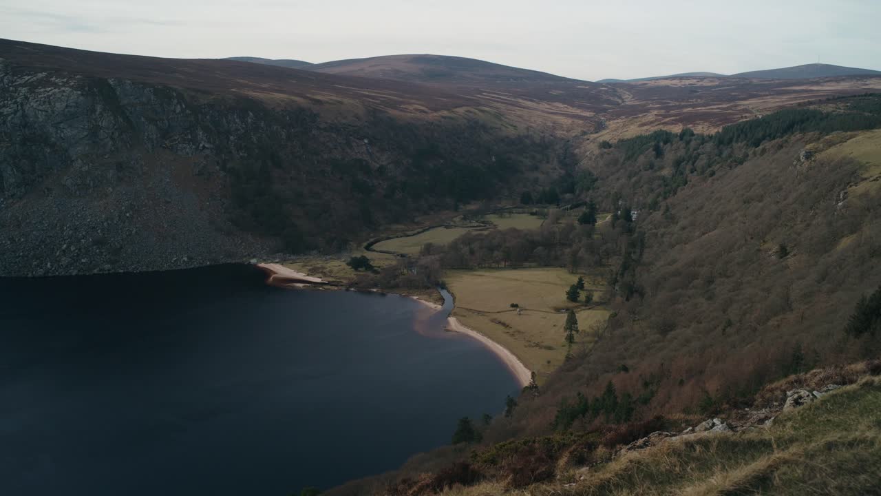 A wide aerial view of Lough Tay in Wicklow, Ireland. The deep blue lake is surrounded by mountains, forests, and rolling hills, offering a breathtaking look at this famous Irish landscape.