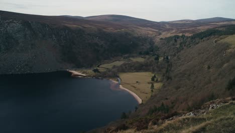 a wide aerial view of lough tay in wicklow, ireland. the deep blue lake is surrounded by mountains, forests, and rolling hills, offering a breathtaking look at this famous irish landscape.