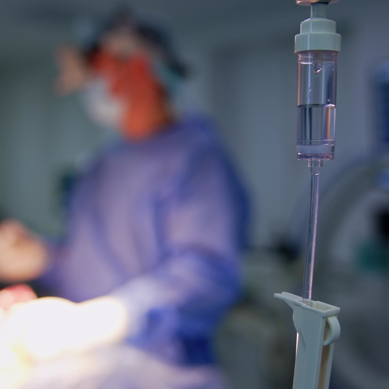 Contatore di gocce sul supporto in sala operatoria. Due medici collaborano durante un'operazione sullo sfondo in modalità sfocata.