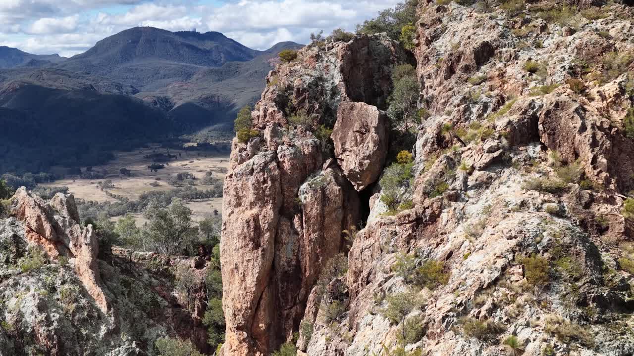 Camera slowly pans across rugged rock formations overlooking forested valleys and distant mountains under bright daylight, highlighting natural textures and dramatic Australian scenery