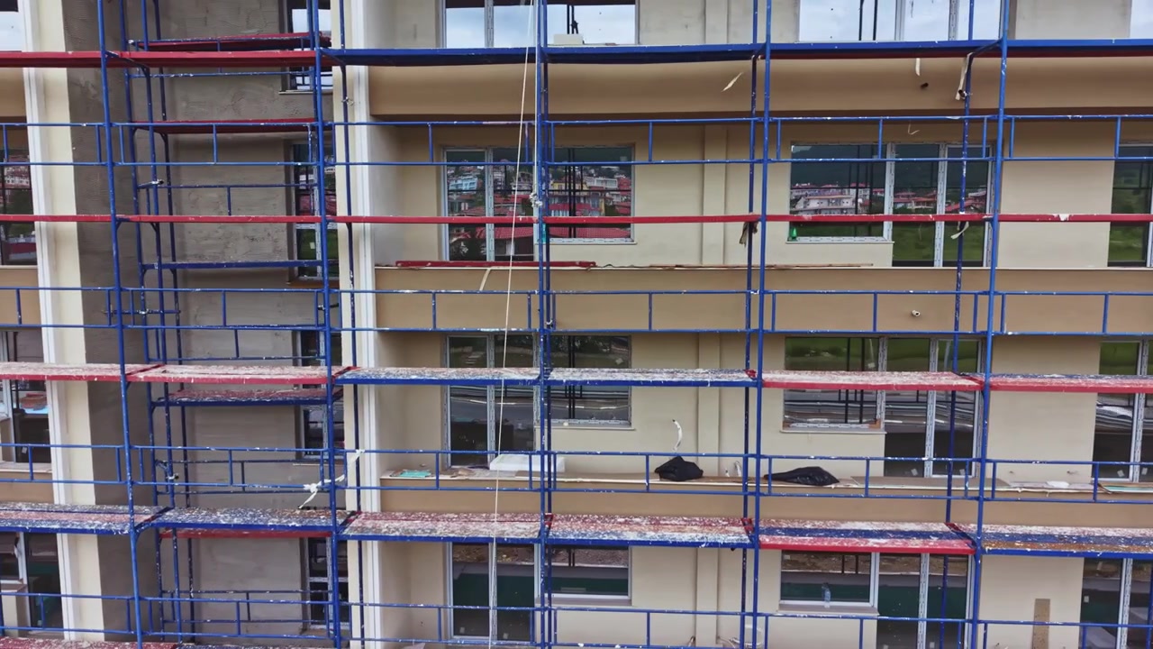 Udvendig konstruktion med synligt stillads. Facaderenovering i dagslys med metalrammer. Folk observerer bygningsrekonstruktion med stillads og beige overflader.