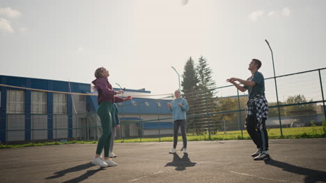 volleyball beginner training outdoors with coach catching ball, surrounded by other players, people walking by behind green bar fence, in sunlit court with building in background