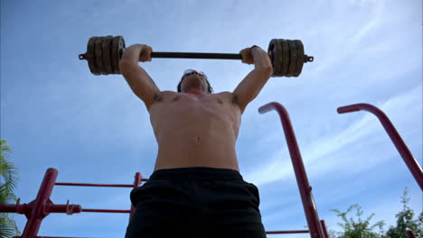 slow motion tilt up shot of an hispanic shirtless man performing several barbell front squat to overhead press at the fitness ground of the park