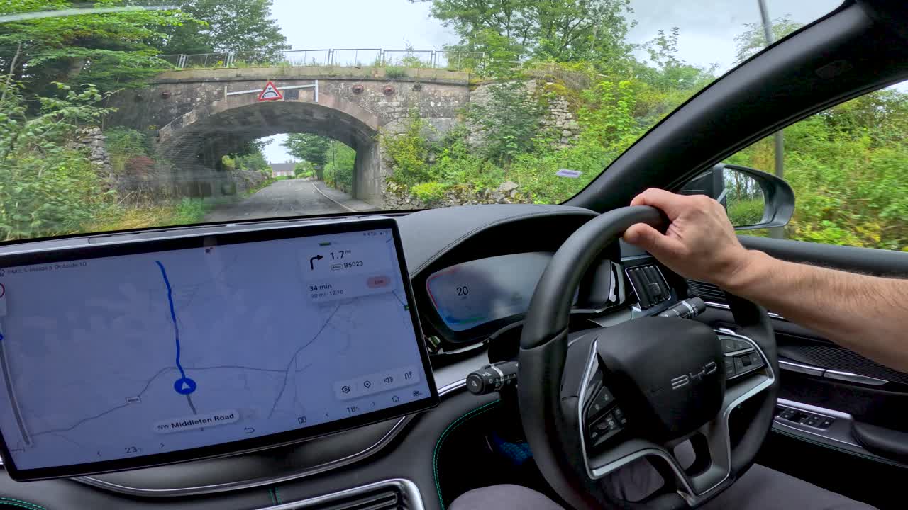 A person drives an electric vehicle along a rural road using a digital navigation system, passing under a historic stone bridge in daylight