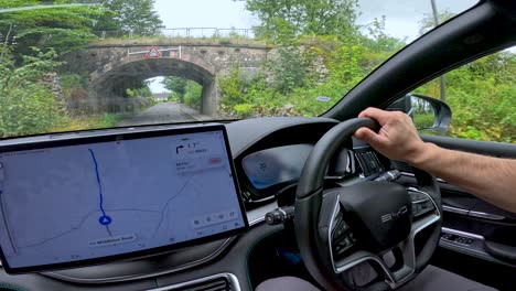 a person drives an electric vehicle along a rural road using a digital navigation system, passing under a historic stone bridge in daylight
