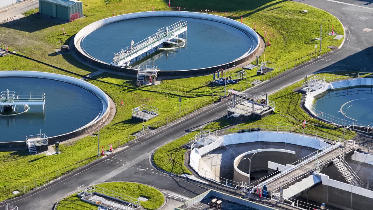 Aerial footage pans across a modern wastewater treatment plant, highlighting circular settling tanks, water channels, and industrial infrastructure in bright daylight