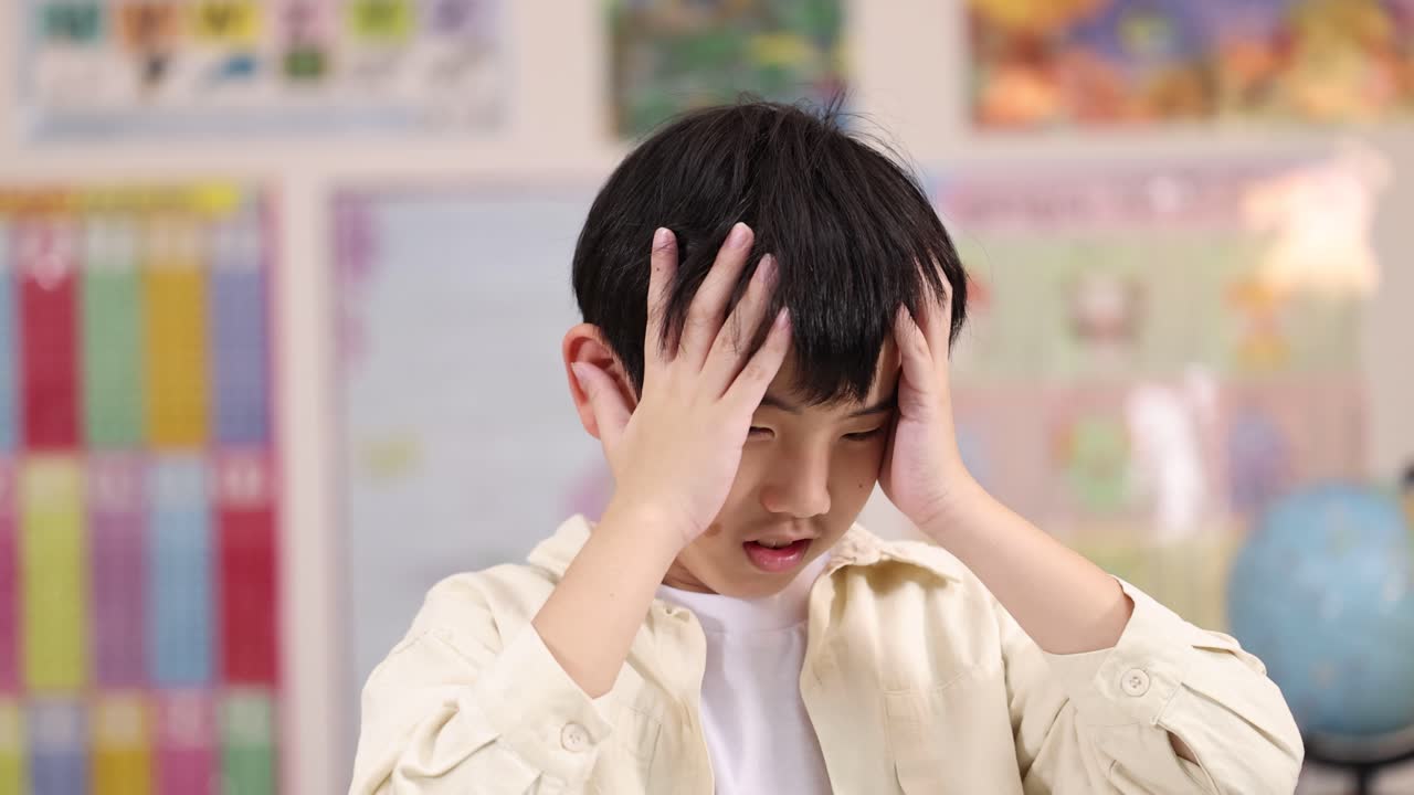 アジア系の男子学生が明るい教室の照明の下で頭を抱え、不満を表している、静的なミディアムショット・コンポジション。