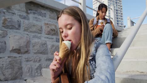 side view of a caucasian and a mixed race girl eating ice cream sitting on stair