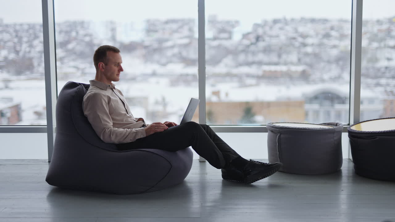 Middle-aged male works on laptop sitting in bean bag chair. Man distracts and looks aside, putting hand to his forehead. Cityscape in blur at backdrop.