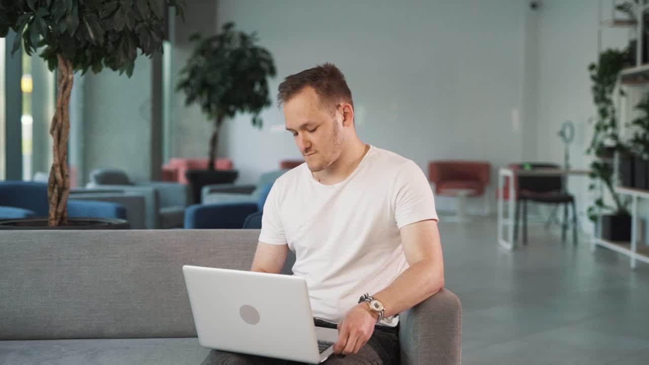 joven en el vestíbulo del centro de negocios se sienta en el sofá, trabaja en su computadora portátil, y refleja