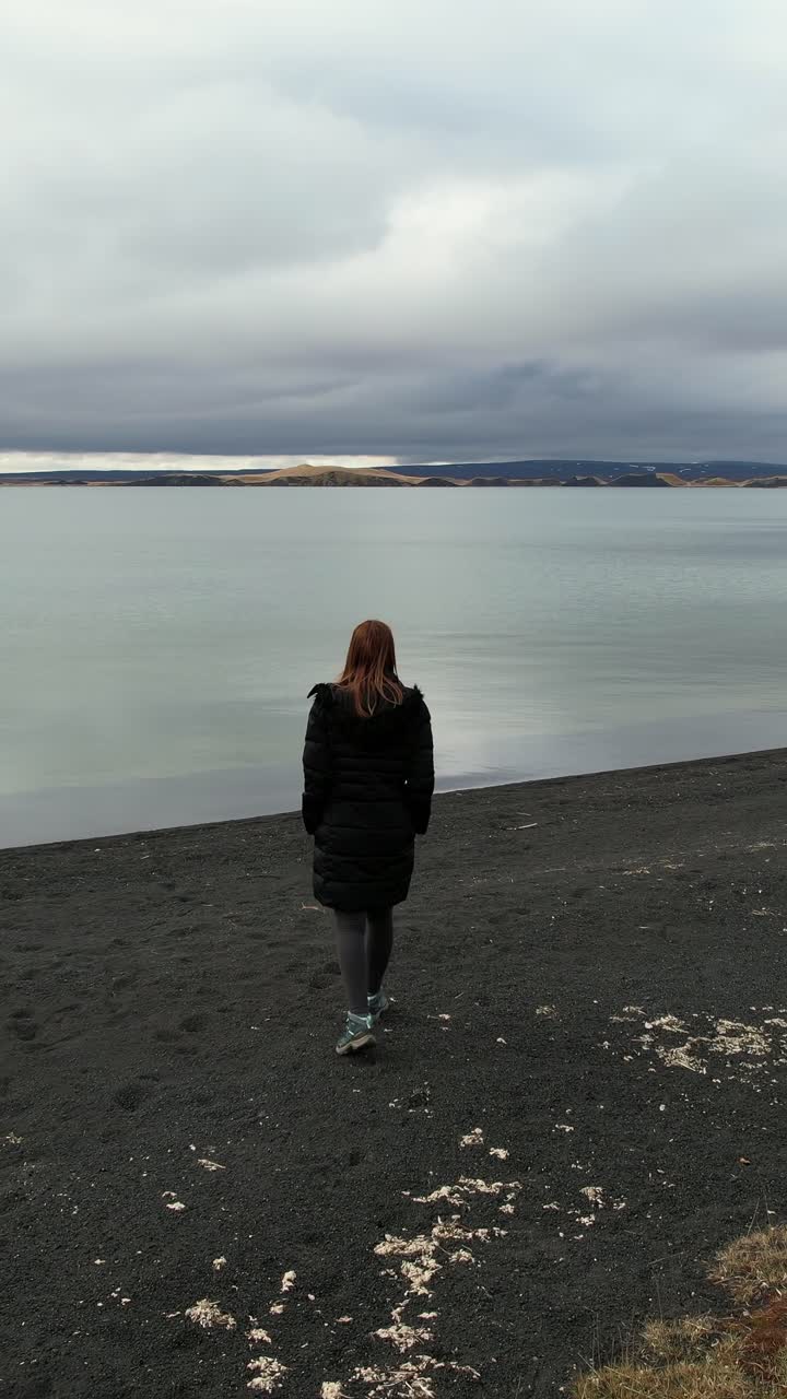Woman walking on a black sand beach towards a lake on a cloudy day in Iceland