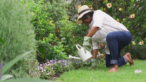 mujer afroamericana mayor con guantes de jardinería regando flores en el jardín