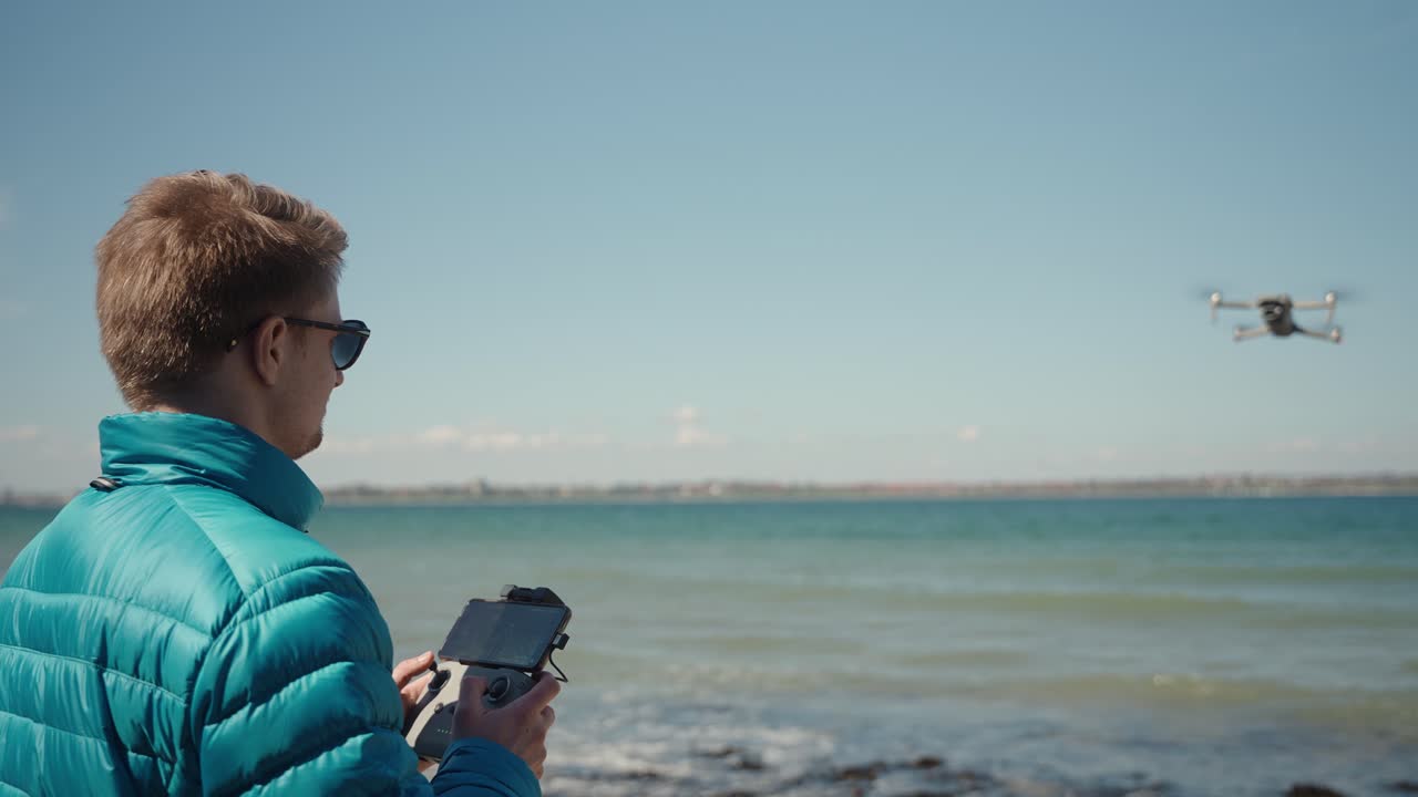 Drone pilot in blue jacket operating drone in front of him while it flies away over the water
