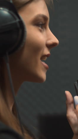 joyful long haired woman vocalist with large headphones sings positive song with microphone in semi-dark modern record studio close side view
