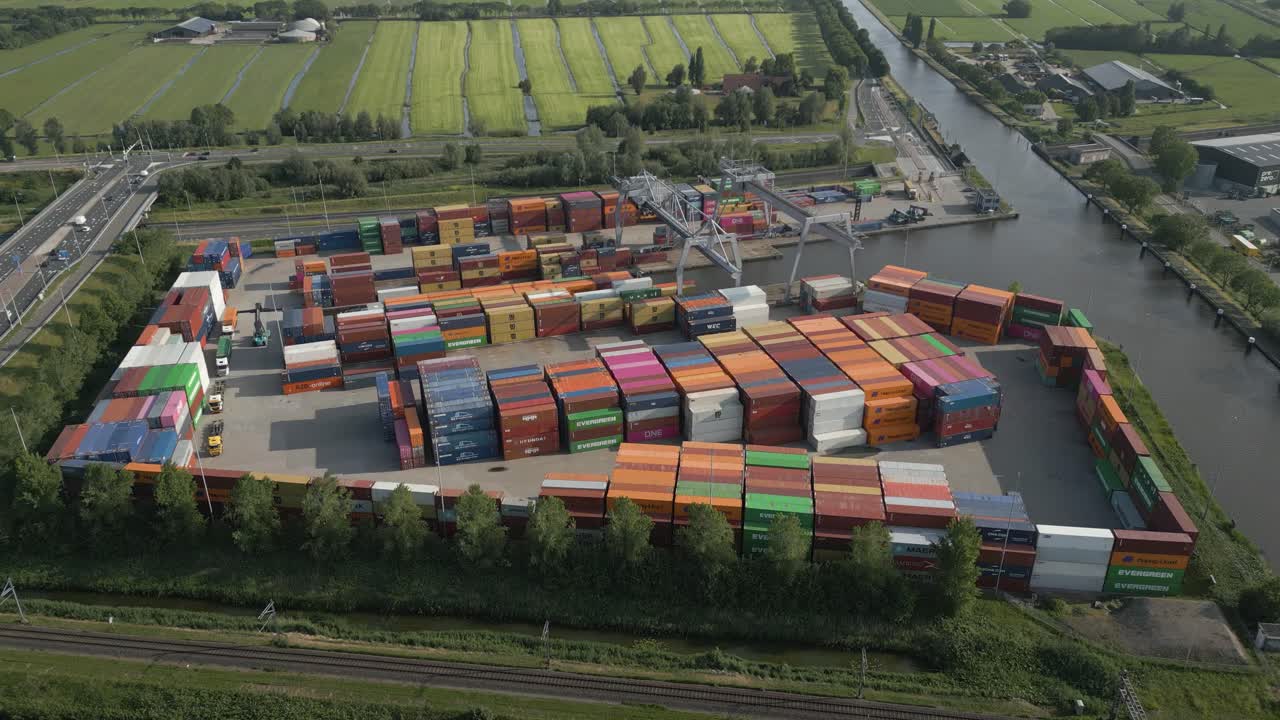Drone shot showing hundreds of shipping containers stacked at a waterside logistics hub near a canal, surrounded by farmland and industry