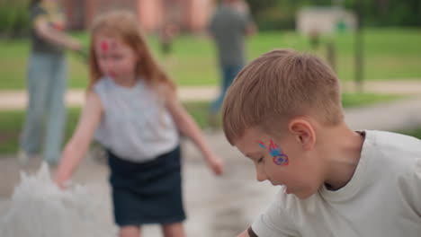 Siblings playing and chatting while touching fountain water, face paint visible, summer park greenery blurred behind, joyful interaction showing curiosity and shared amusement