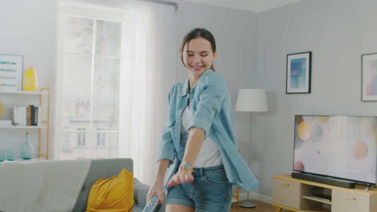 Slow Motion Shot of a Young Beautiful Woman in Jeans Shirt and Shorts Dancing and Vacuum Cleaning a Carpet in a Cozy Room at Home. She Uses a Modern Cordless Vacuum. She's Happy.
