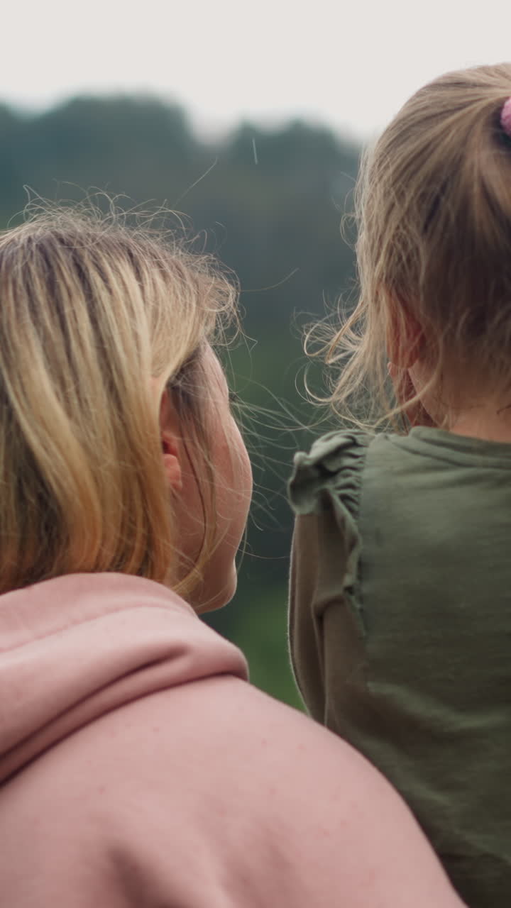 Happy woman hugs little children talking to daughter while looking at hills covered with forest on cloudy rainy cool day backside view