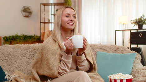 Adult woman wrapped in blanket sitting on sofa with cup of tea watching series thoughtfully at home