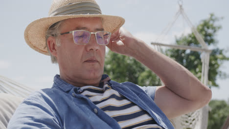 Happy-senior-caucasian-man-in-sunhat-sitting-in-hammock-using-tablet-on-beach,-in-slow-motion