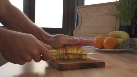 Hombre-Cortando-Pan-De-Croissant-Con-Un-Cuchillo-En-Una-Tabla-De-Madera-En-La-Cocina-Para-Desayunar