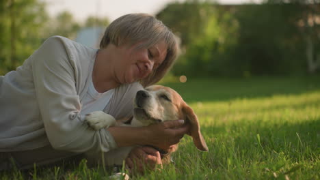 primer plano de una mujer acostada en un campo de hierba sosteniendo a su perro con afecto mientras acaricia su cara, bajo la cálida luz del sol, con un fondo borroso de árboles y vegetación