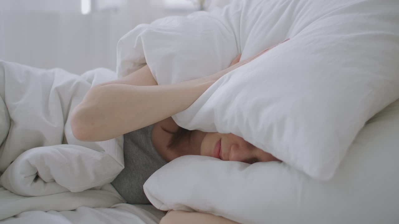 짜증나는 여성은  ⁇ 선 소음으로 아침 일찍 깨어났습니다. 여성은 아침 일찍 침실에서 소음을 듣지 않기 위해 베개로 자신을  ⁇ 고 있습니다.