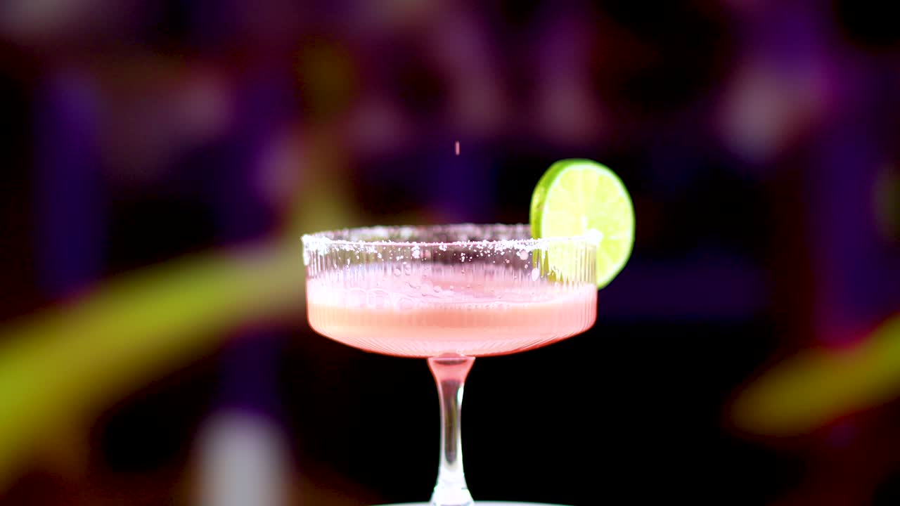A pink margarita is poured into a glass, garnished with a lime slice, against a colorful background