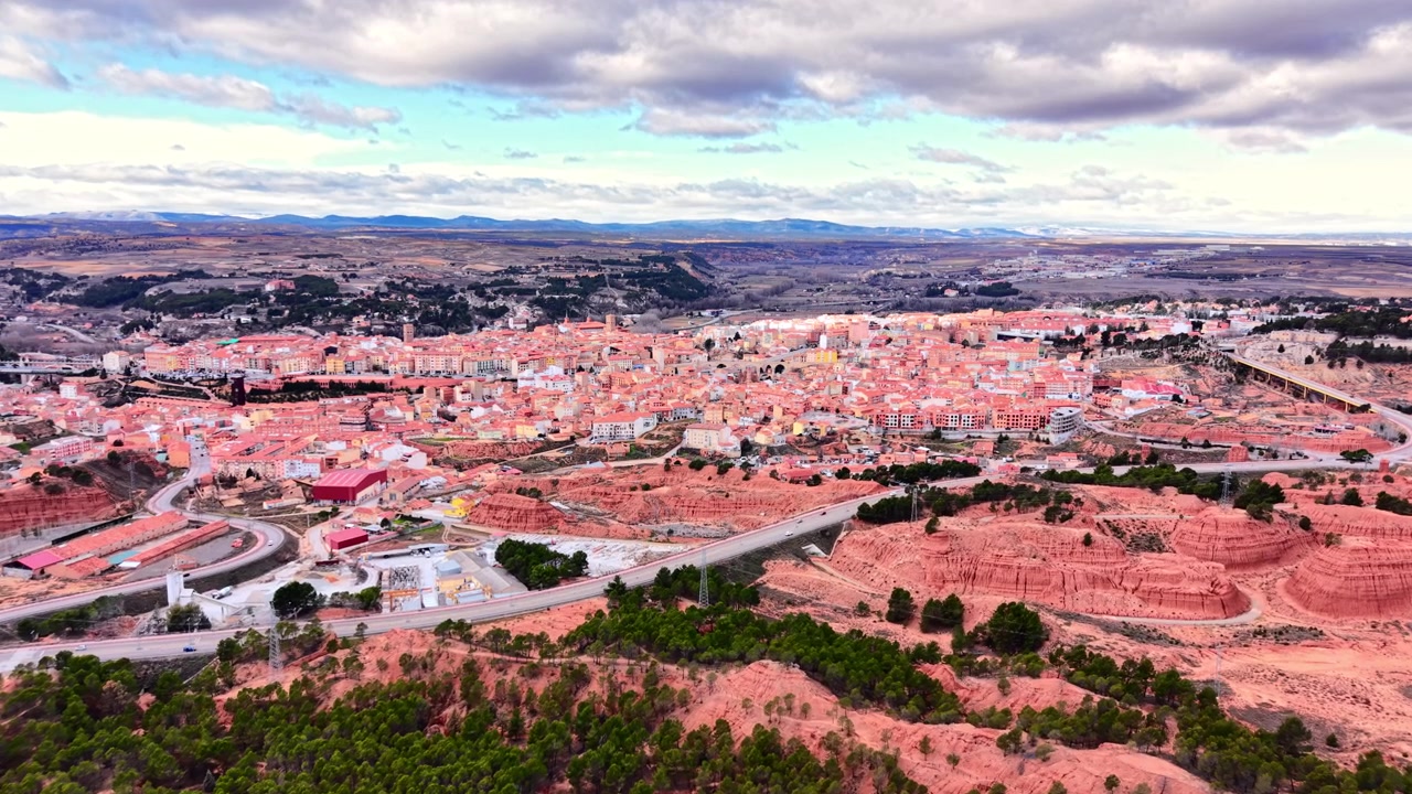 スペイン、テルエルの赤い岩の悪地帯に囲まれた丘の上の町のパノラマビュー。テラコッタの屋根の都市集落、松の木、浸食された赤い砂岩の地形、谷、そして曇り空の山脈が見える。