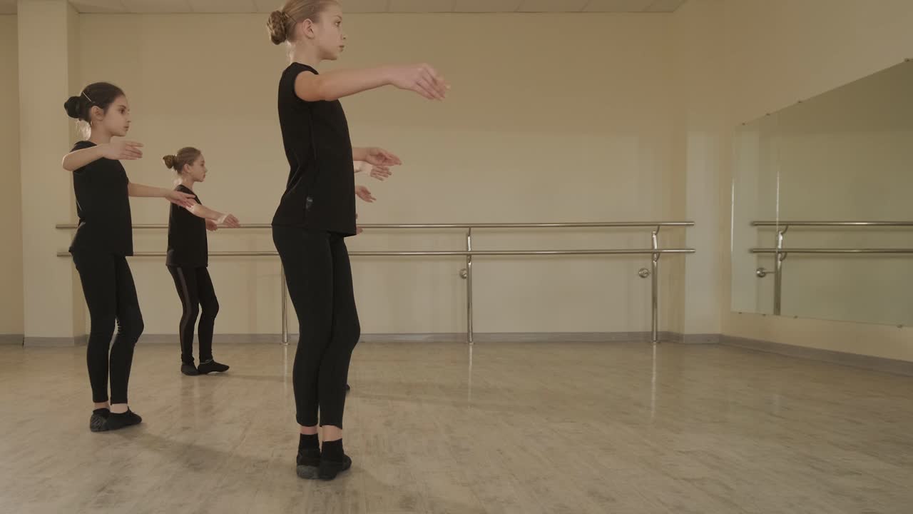 un grupo de jóvenes estudiantes de ballet con ropa de baile negra practicando posiciones en un amplio estudio de ballet con pisos de madera y barras montadas en la pared. expresiones enfocadas y movimientos sincronizados.