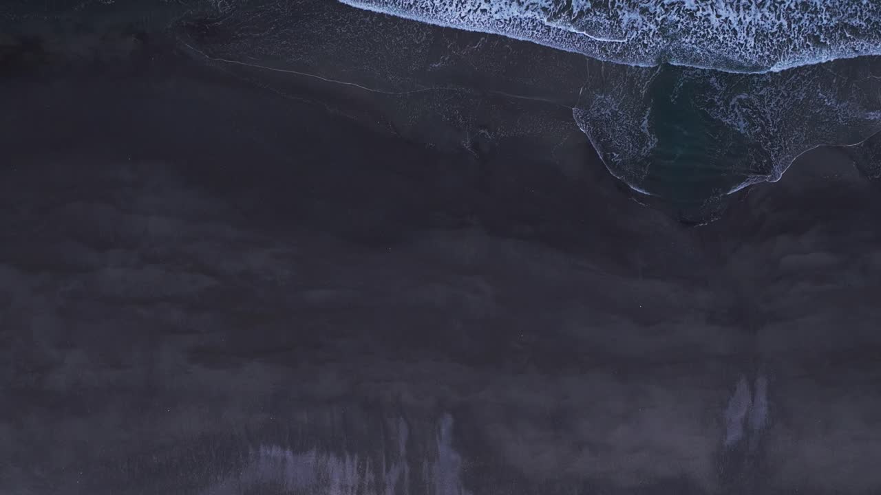 Drone shot looking straight down as it flies over waves crashing on the beach