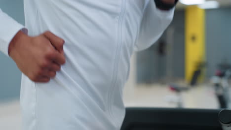 close up partial view of athlete in white top running on treadmill in modern gym, arm and torso in frame, blurred equipment and yellow accent wall in background