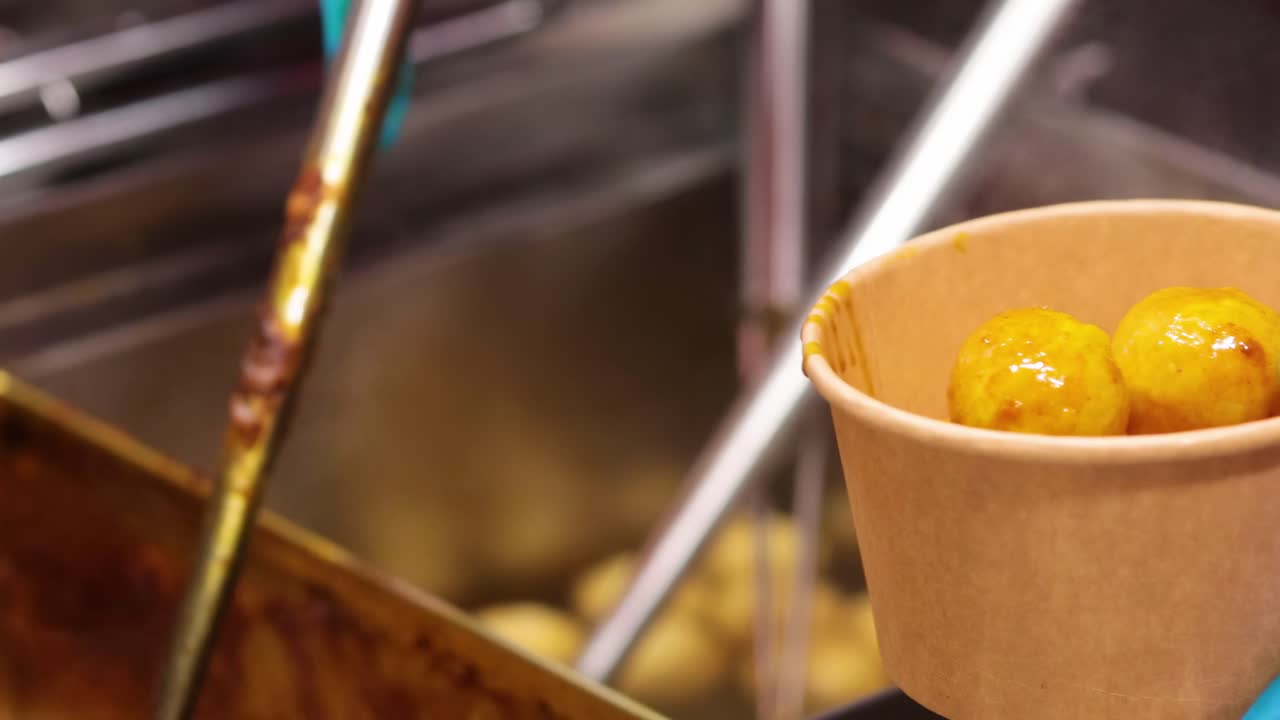 A close-up of blue-gloved hands placing steaming meatballs into a cardboard bowl using a perforated ladle.