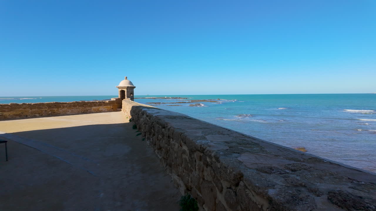 A coastal promenade in C&aacute;diz featuring a small sentry box at the end of a stone wall, overlooking the Atlantic Ocean under a vast blue sky